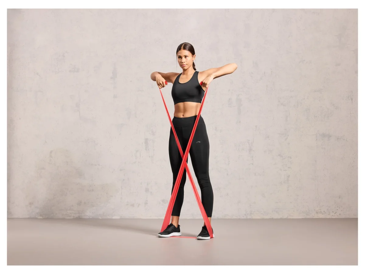 Donna allena braccia con fascia elastica rossa Crivit incrociata sotto i piedi durante allenamento